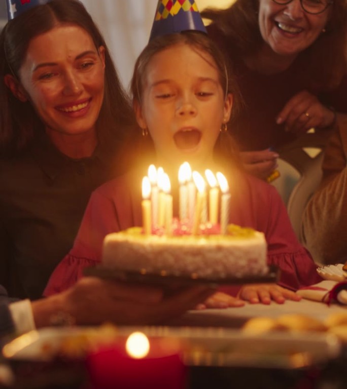 垂直画面:快乐兴奋的女孩吹灭美味生日蛋糕上的蜡烛。年轻快乐的孩子被她的家人、亲戚和朋友包围着，坐在舒