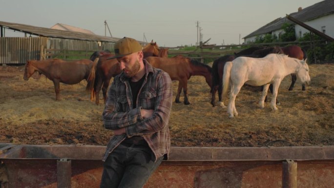 一个年轻的农场工人的肖像，背景是篱笆和马。一个年轻的农民在马厩里照看马。
