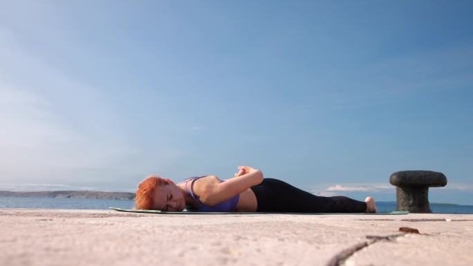 阳光下躺在海边瑜伽垫上的中年妇女，双手放在背后，抬起上半身