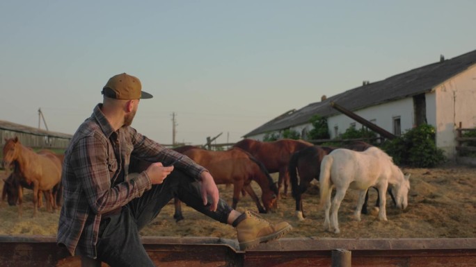 一个年轻人手里拿着手机，背景是一个有马的围场。一个人在农场工作了一天后休息，夕阳西下。