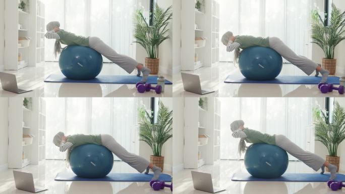 运动的伊斯兰妇女穿着穆斯林运动服，打普拉提运动球，在瑜伽垫上锻炼肩部肌肉，在家里客厅的笔记本电脑上进