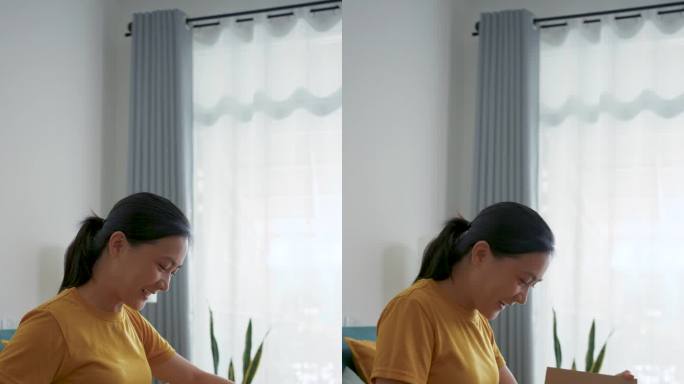 一名亚洲妇女坐在家里的客厅里，打开网上购物的纸箱，打开包装，检查商品。