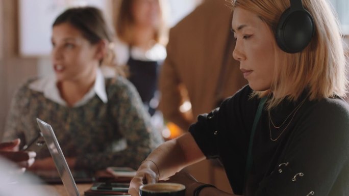 年轻的亚洲女性用笔记本电脑在咖啡馆工作，打字，在社交媒体上分享信息，戴着耳机，在繁忙的餐馆里享受听音