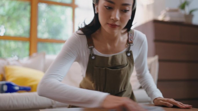 亚洲美女在家打扫室内客厅，女孩家庭主妇管家清洁工感到快乐和擦拭凌乱肮脏的工作台面为管家家务或杂务。