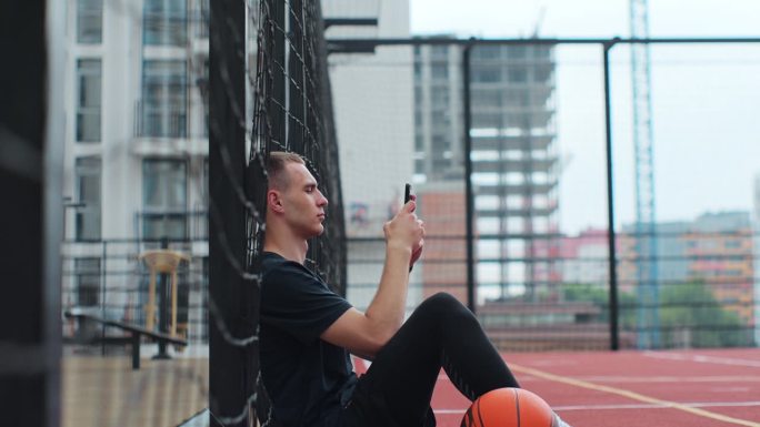 侧视图，年轻的运动男子坐在篮球场上，滚动智能手机，使用应用程序。在户外运动场上休息。爱好、运动、生活