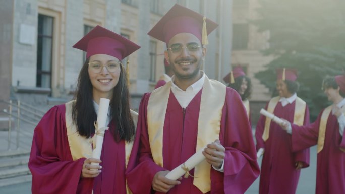 戴着毕业帽、身穿毕业服的白人女性和阿拉伯男性站在校园里，手里拿着毕业证书，面带微笑