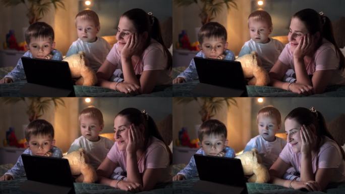 微笑的年轻母亲带着两个儿子躺在床上用平板电脑看电影。家人聚在一起，养育孩子，看视频，上床睡觉
