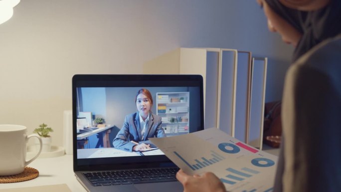 年轻快乐的亚洲穆斯林女秘书戴着头巾，穿着休闲服，视频会议，专注于笔记本电脑屏幕，晚上在家里与女商人加