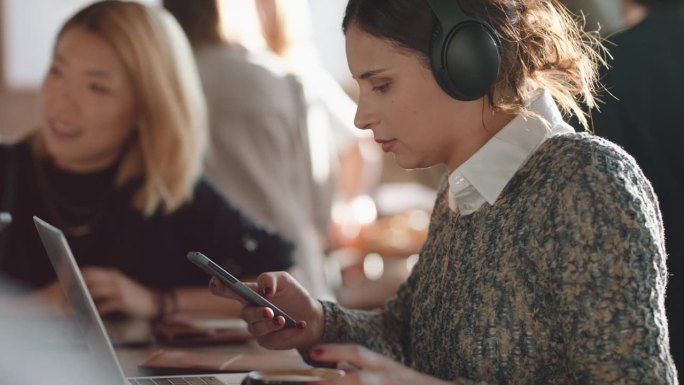 年轻女子用笔记本电脑在咖啡馆工作，打字，在社交媒体上分享信息，戴着耳机，在繁忙的餐馆喝咖啡，享受听音