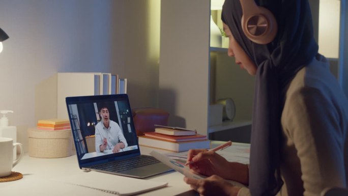 年轻快乐的亚洲穆斯林女秘书戴着头巾，穿着休闲服，在笔记本电脑上与商人在家里进行视频会议，晚上加班。小