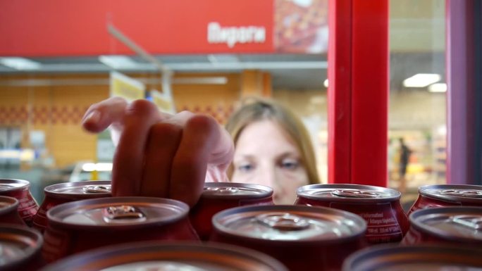 一个商店的冰箱里有很多铝罐苏打水，一个年轻女子打开门，拿了一个