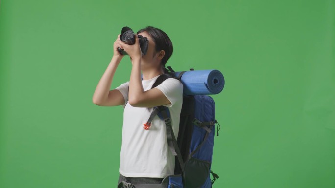 亚洲女徒步者背着登山背包站在绿幕背景上用相机拍照的侧视图