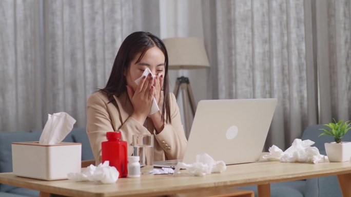 生病的亚洲女商人在家里的客厅用笔记本电脑工作。生病的女孩打喷嚏，用纸巾擦鼻涕