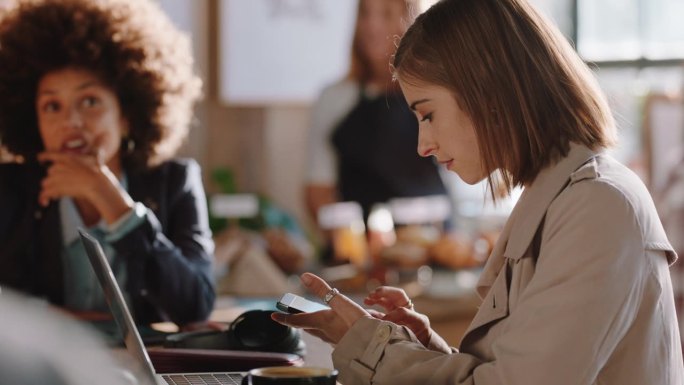 年轻女子用笔记本电脑在咖啡馆工作，在智能手机上打字，在社交媒体上分享信息，享受坐在繁忙的餐厅里
