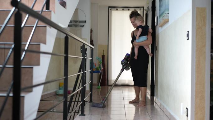 母亲抱着女婴一边打扫房子，一边用吸尘器打扫走廊