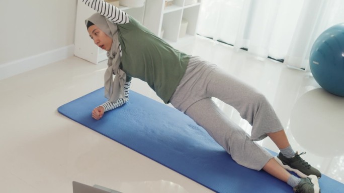 运动亚洲伊斯兰妇女穿着穆斯林运动服锻炼伸展腿和流动手臂初学者练习在线课程训练从笔记本电脑在家里的客厅