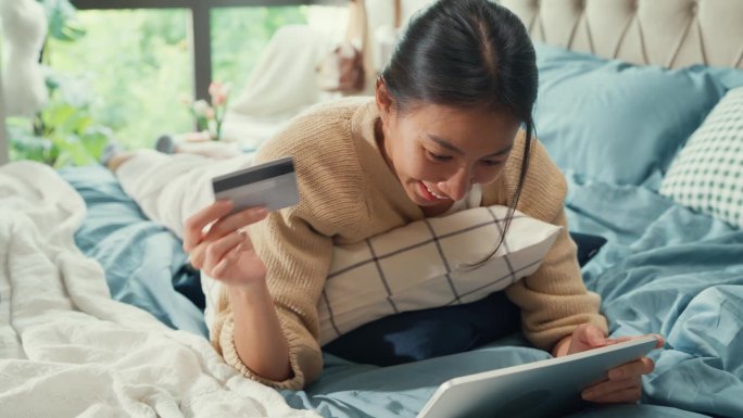 快乐微笑的亚洲女子大学白色奶油睡衣躺在柔软舒适的床上使用信用卡网上购物成功支付在卧室在家里的数码平板