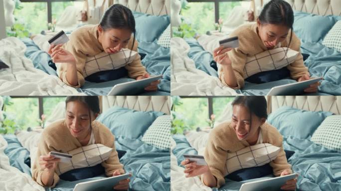 快乐微笑的亚洲女子大学白色奶油睡衣躺在柔软舒适的床上使用信用卡网上购物成功支付在卧室在家里的数码平板