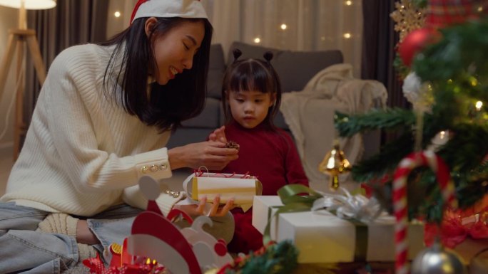 亚洲年轻快乐的家庭晚上在家里一起享受新年假期聚会。妈妈和孩子一起把礼品盒放在圣诞树旁装饰。度假的生活