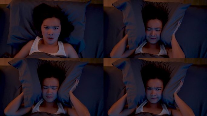 亚洲女性对噪音感到厌烦，失眠，躺在卧室的床上捂着耳朵。