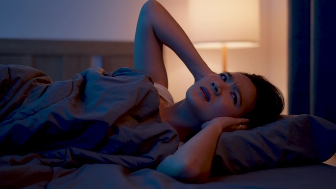 亚洲女性对噪音感到厌烦，失眠，躺在卧室的床上捂着耳朵。