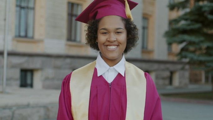 大学校园里，戴着毕业帽，穿着毕业礼服的快乐的非裔美国妇女的慢动作肖像