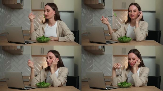 一位有思想的年轻女子坐在厨房里使用笔记本电脑，吃了一份沙拉，感觉没有胃口。消化问题，以及变质和无味的