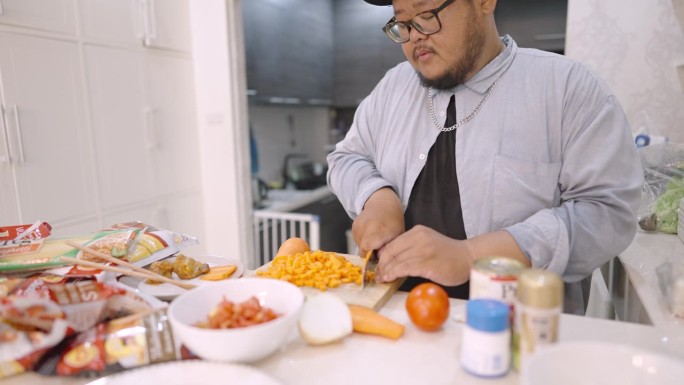 一个肥胖的男人一边欢快地唱着歌，一边在厨房里把胡萝卜切成小方块，放上一大堆食物，准备在家做饭。