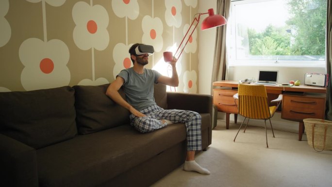 年轻人戴着虚拟现实耳机坐在家里的沙发上观看360度视频想象概念