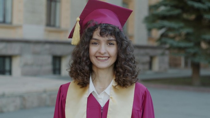 穿着毕业袍，戴着毕业帽，自信的年轻女子站在校园里微笑，感到自豪的慢动作肖像