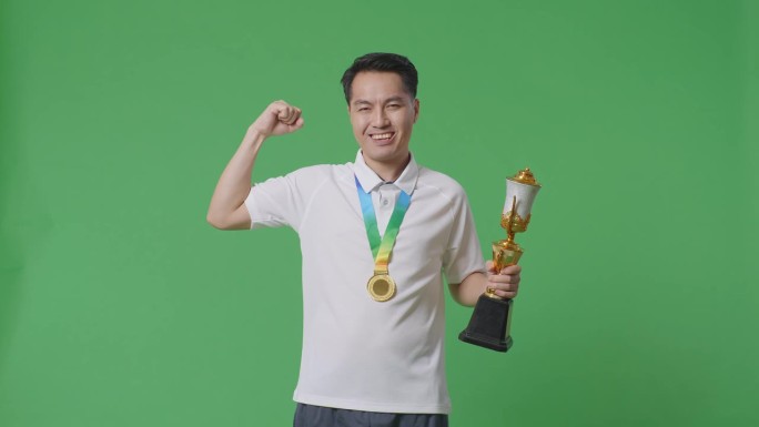 一名亚洲男子拿着金牌和奖杯，在摄影棚的绿幕背景下，对着镜头微笑，展示着他的二头肌