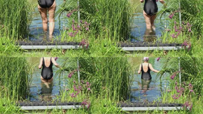 一个长着脂肪团的成年女子，穿着黑色连体泳衣，慢慢地走进清澈透明的湖水中游泳