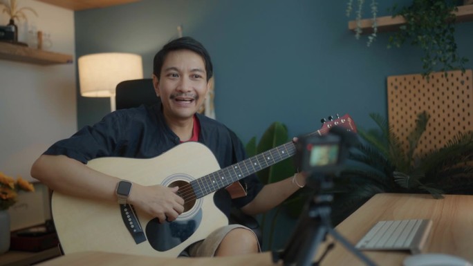 有吸引力的亚洲人有才华的艺术家录制视频的智能手机坐在桌子上在现代工作室弹吉他。