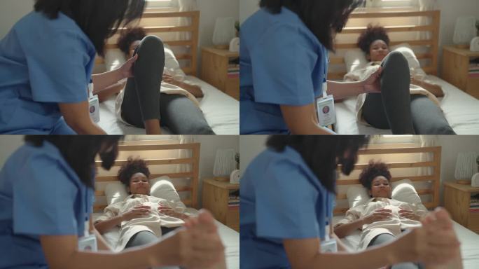 出诊期间，保健医生女来访者和非裔美国孕妇躺在病床上。好心的亚洲医生或理疗师帮助和支持年轻孕妇在家做腿