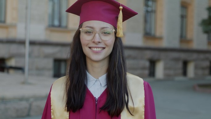 戴着毕业帽、穿着毕业礼服的聪明女孩在校园里微笑的慢动作肖像