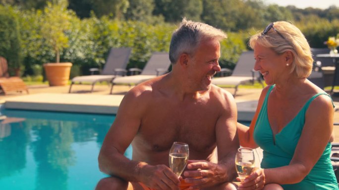度假的老夫妇坐在躺椅上，在酒店游泳池边用香槟烤面包