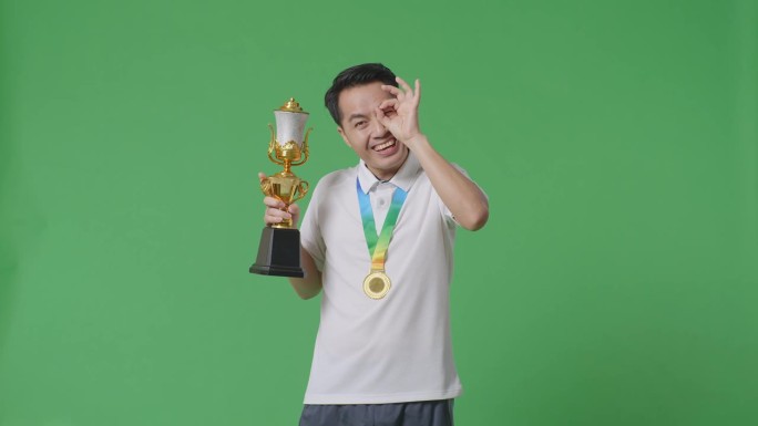 一名亚洲男子拿着金牌和奖杯，在绿幕背景下对着镜头微笑，他是第一个获得金牌的人