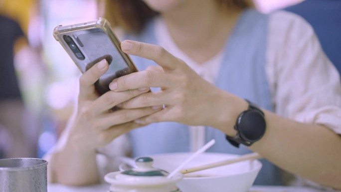 在一家面馆，一名亚洲女性在午休时间一边用筷子喝着一碗面汤，一边用智能手机阅读在线信息。