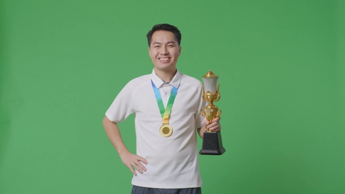 一名亚洲男子拿着金牌和奖杯对着镜头微笑，单臂叉腰，在演播室的绿幕背景下摆出第一名获胜者的姿势