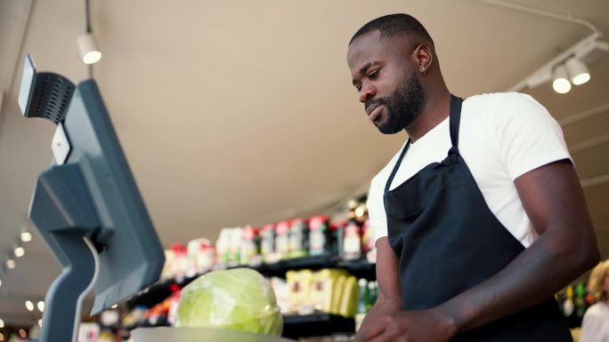 一个穿着白t恤、系着黑色围裙的黑皮肤男子在超市用电子秤称蔬菜的重量