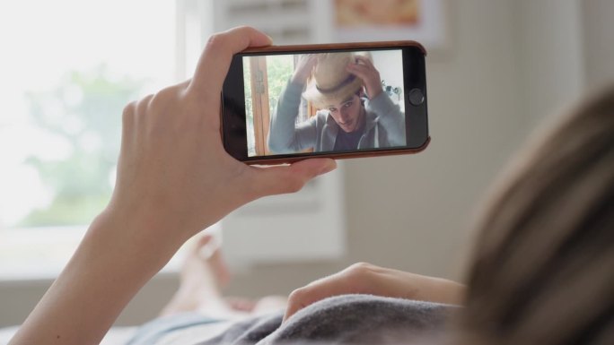年轻女子拿着智能手机摄像头和躺在家里床上的朋友进行视频聊天