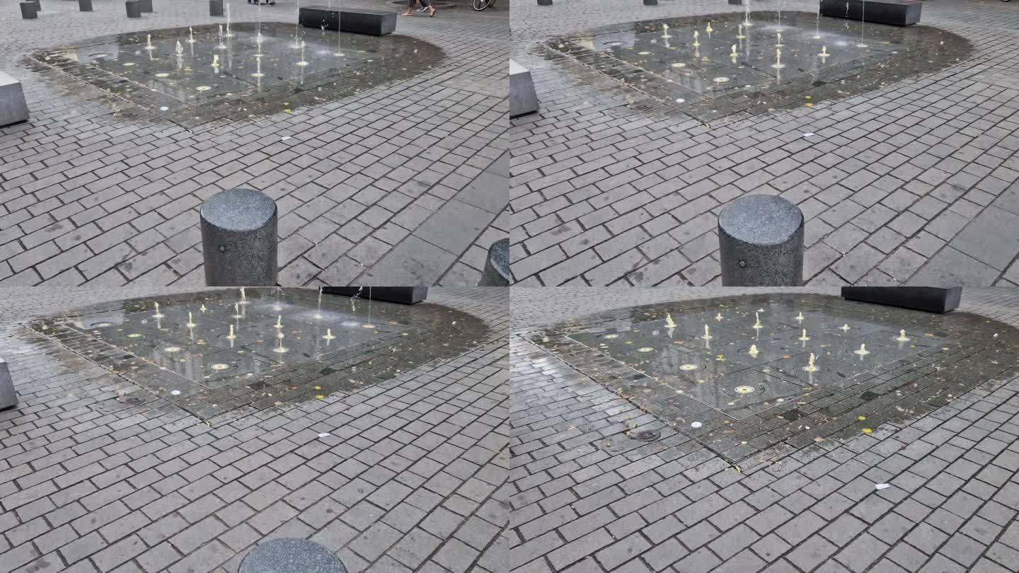 方形花岗石铺装在人行道上一条条地堆放着，水花以交替的高度喷射。水景直接出现在公园街的鹅卵石广场上。湿