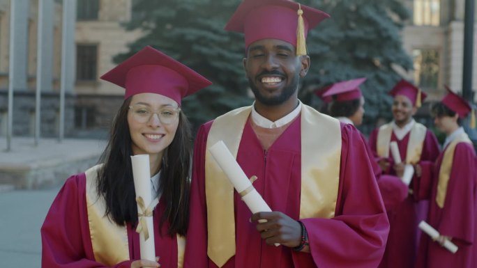 快乐的毕业生，女孩和男孩站在校园里，穿着长袍，戴着帽子，拿着毕业证，享受毕业的日子