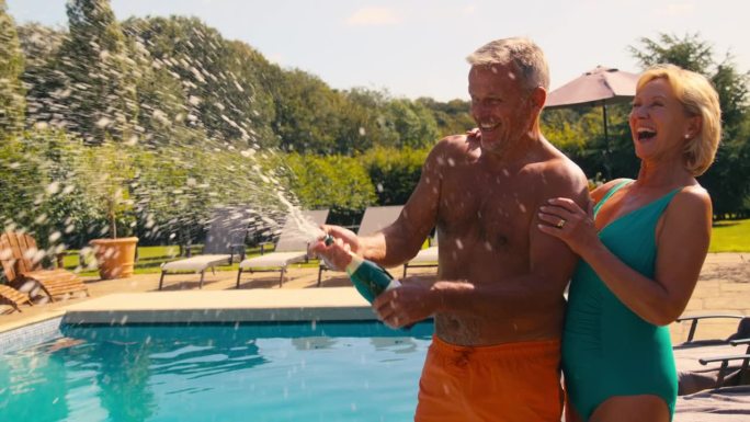 穿着泳装度假的老夫妇在泳池边开瓶喷香槟酒