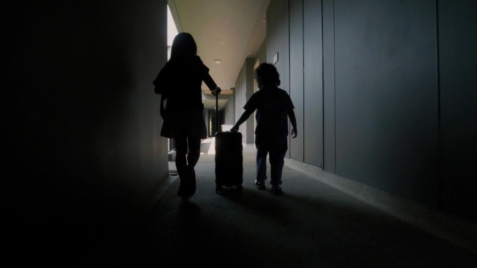 一个亚洲小女孩和她的弟弟走在酒店的走廊上，手里拿着旅行行李。