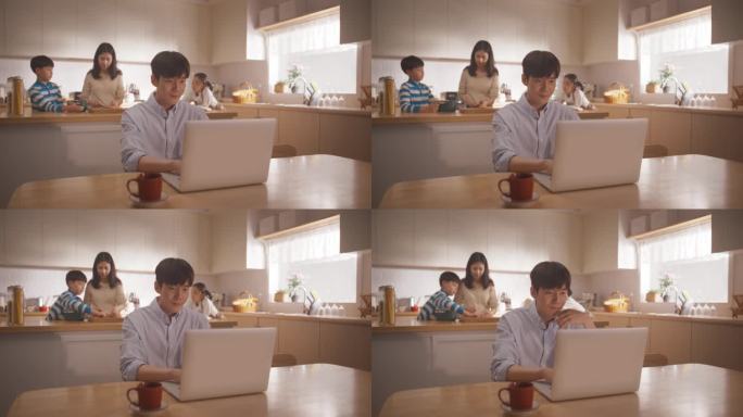 年轻的韩国父亲使用笔记本电脑在家工作，而他的妻子和孩子正在一起准备午餐。忙碌的商人在家办公和联系同事