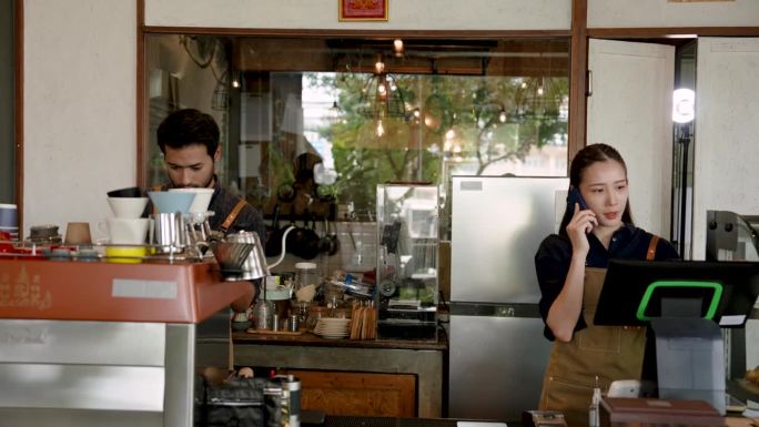 夫妻俩经营着一家小咖啡店，亚洲女人接听老顾客的电话，男咖啡师在柜台后面为顾客准备咖啡。