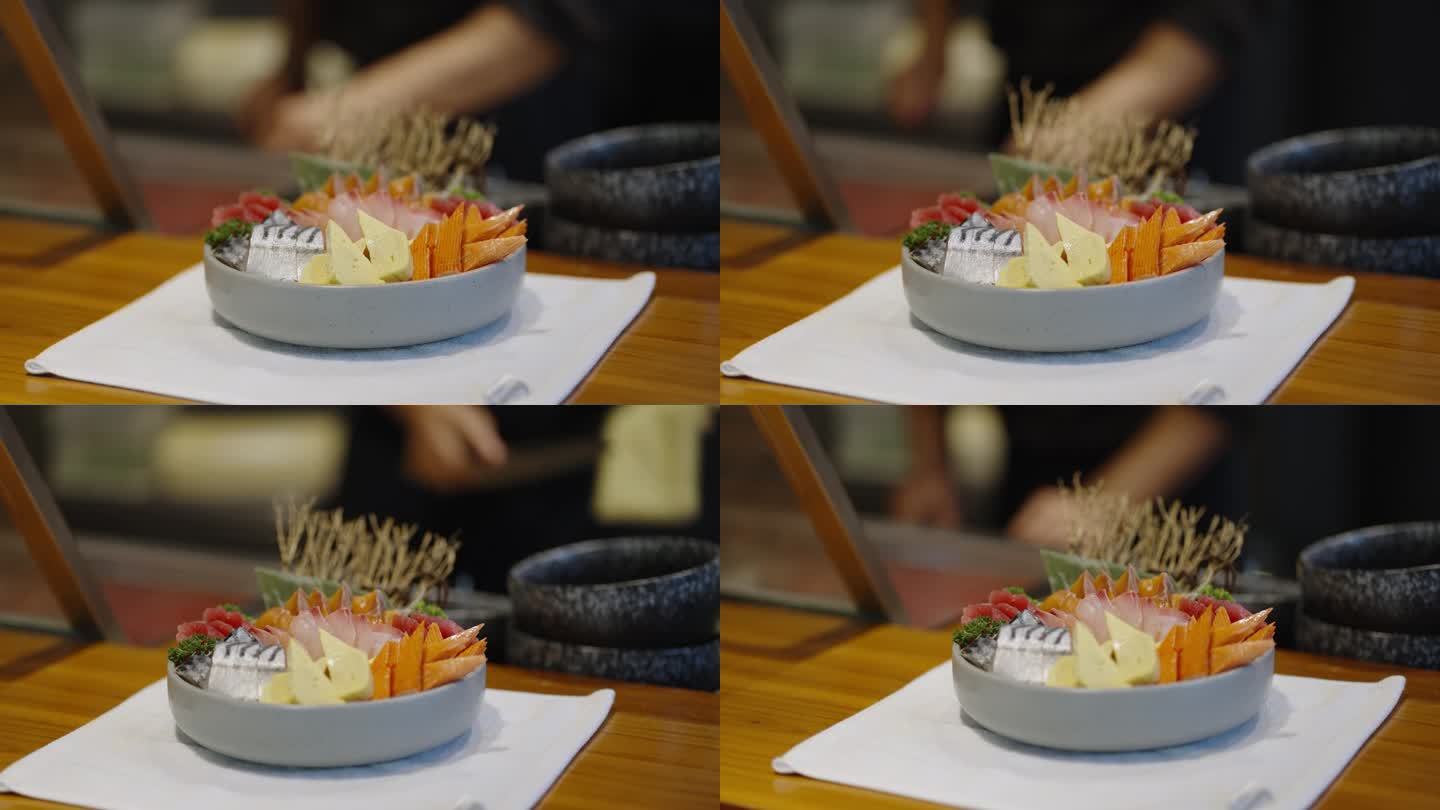 寿司吧柜台上装饰精美的彩色鱼和海鲜刺身盘特写。背景是寿司师傅在厨房吧台后面切生鱼和用毛巾擦菜刀的模糊