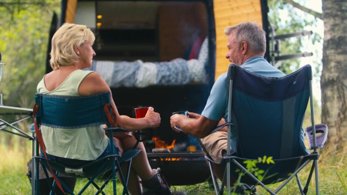 一对老年夫妇开着房车在乡村露营的后景，他们在户外的火炉旁喝咖啡