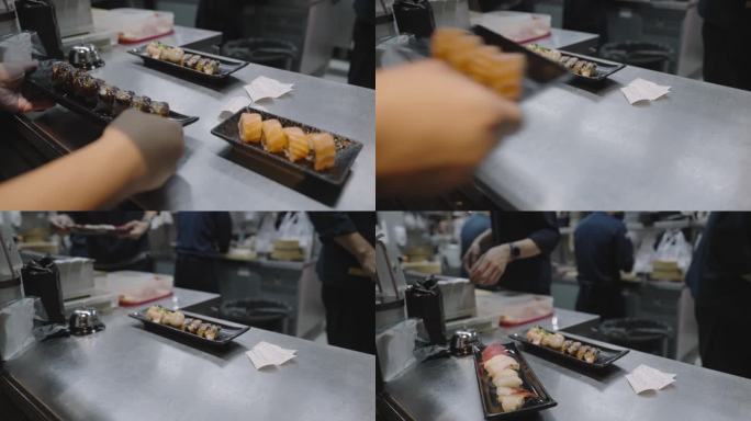 在一家寿司店，一名服务员根据订单在厨房柜台上拿起寿司盘，而一小群厨师正在准备和烹饪日本料理。
